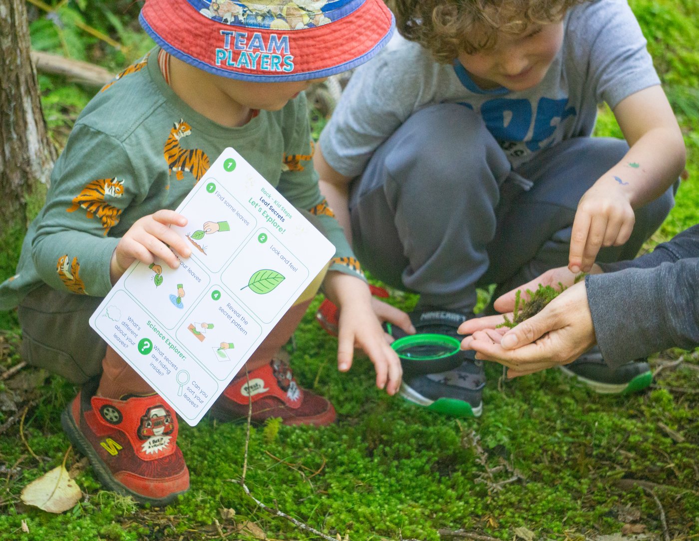 Two young children exploring the bush floor together, one holding a Wild Wonders activity card while an adult’s hands hold a piece of moss